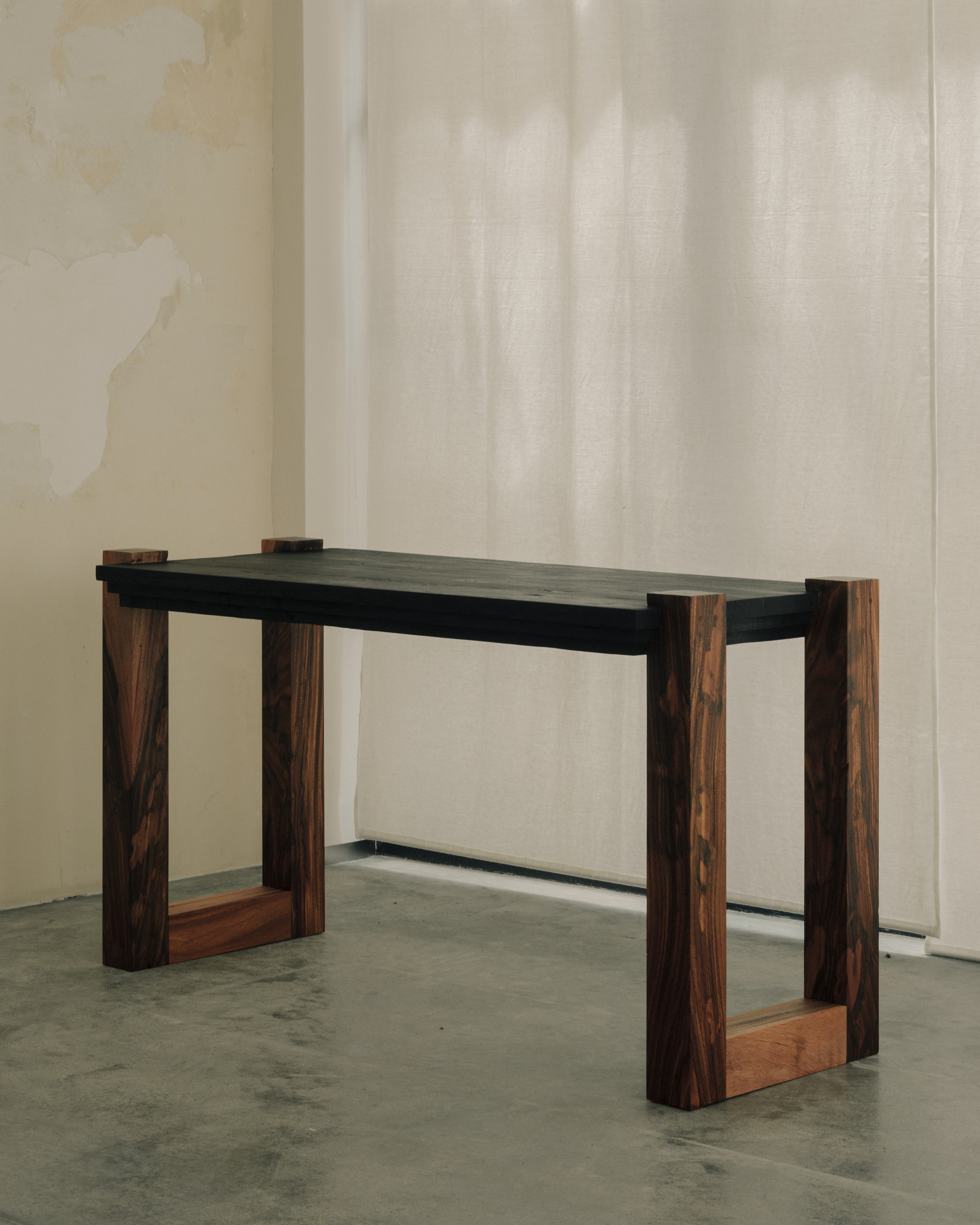 The writing desk shown at an angle in a bright room with a linen curtain and concrete floor